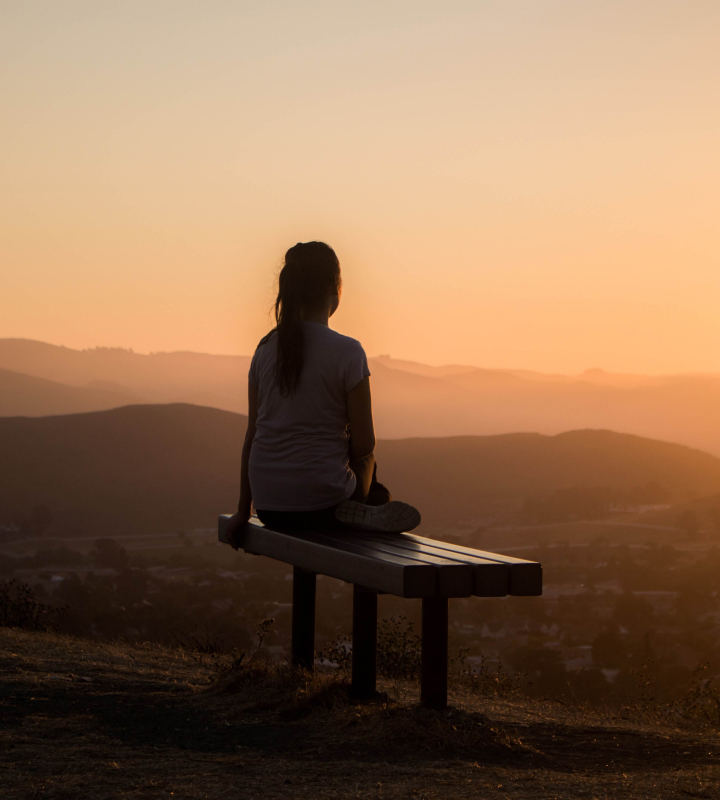 Menina sentada em banco observa o pôr do sol.